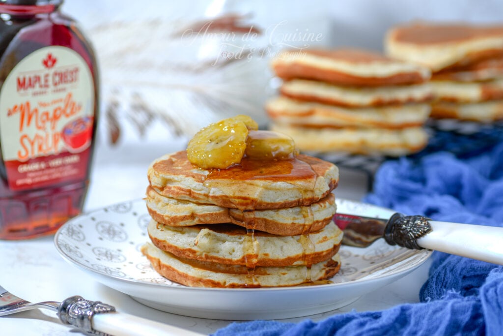 Stack of banana pancakes on a plate, drizzled with maple syrup and topped with banana slices; a syrup bottle sits on the left, with more pancakes on a cooling rack in the background and a blue cloth nearby.