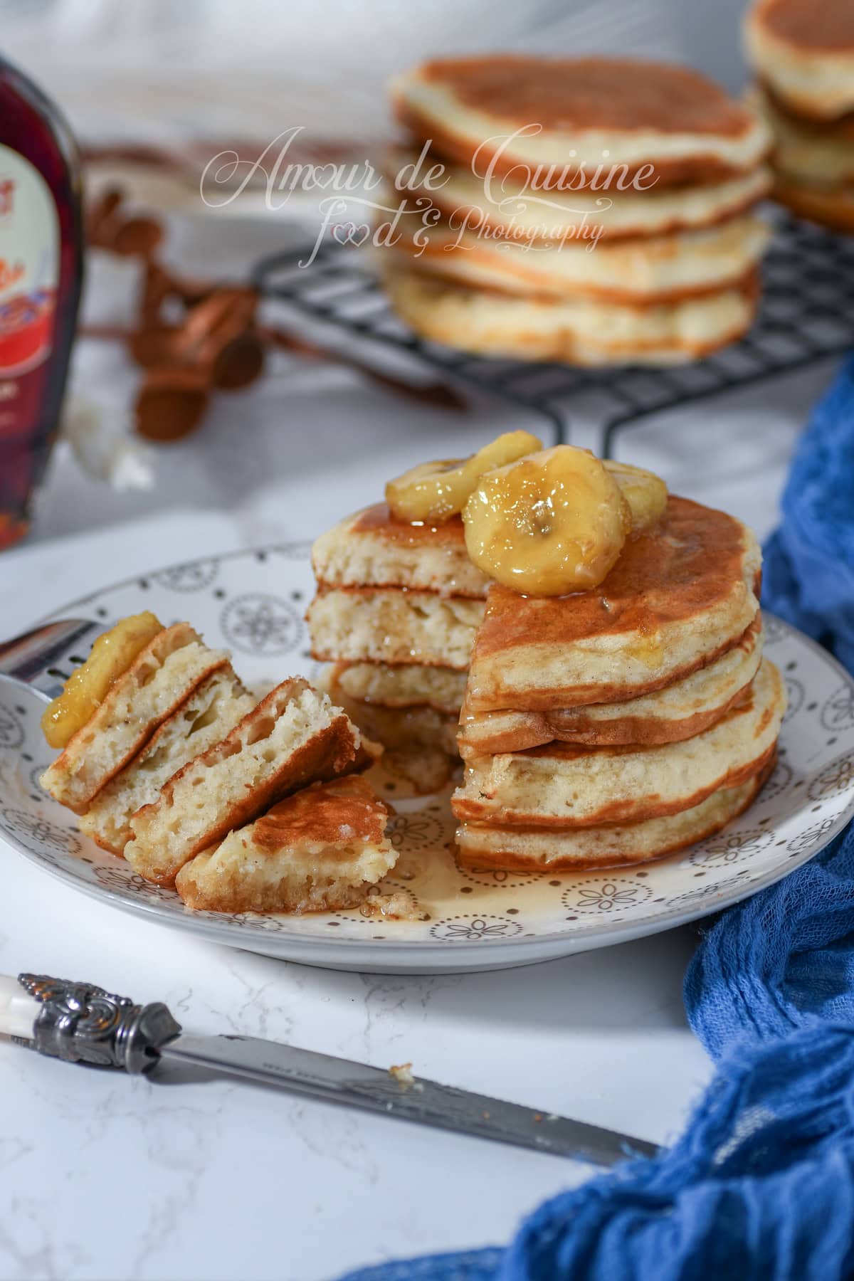 Golden banana pancakes stacked on a plate, drizzled with syrup and topped with banana slices; one pancake is cut open to show a fluffy crumb, with a cooling rack and more pancakes softly blurred in the background.