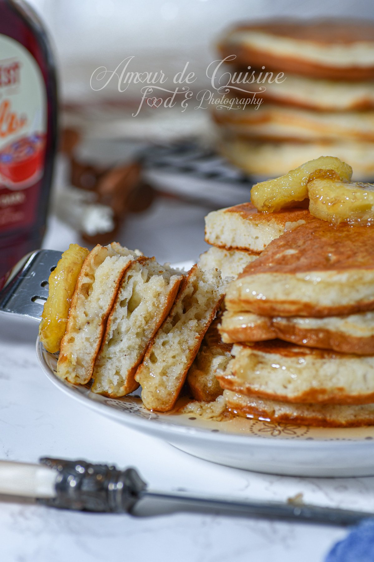 Close-up of banana pancakes: a golden stack drizzled with syrup and topped with banana slices; sliced pieces reveal a fluffy, airy crumb, with a syrup bottle and a cooling rack softly blurred in the background.