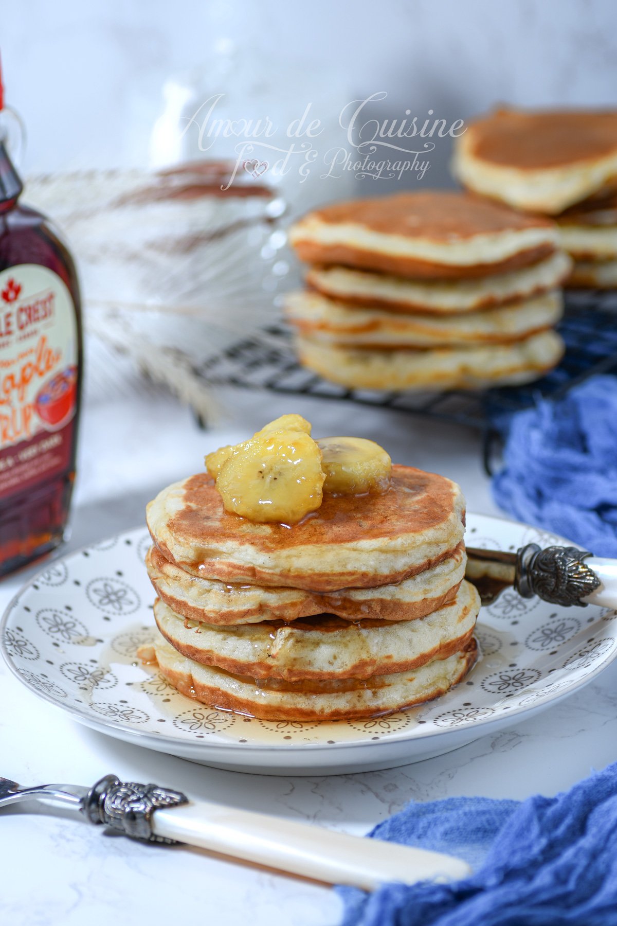 Stack of banana pancakes on a patterned plate, drizzled with maple syrup and topped with banana slices; a syrup bottle on the left, and more pancakes on a cooling rack in the background with a blue cloth.