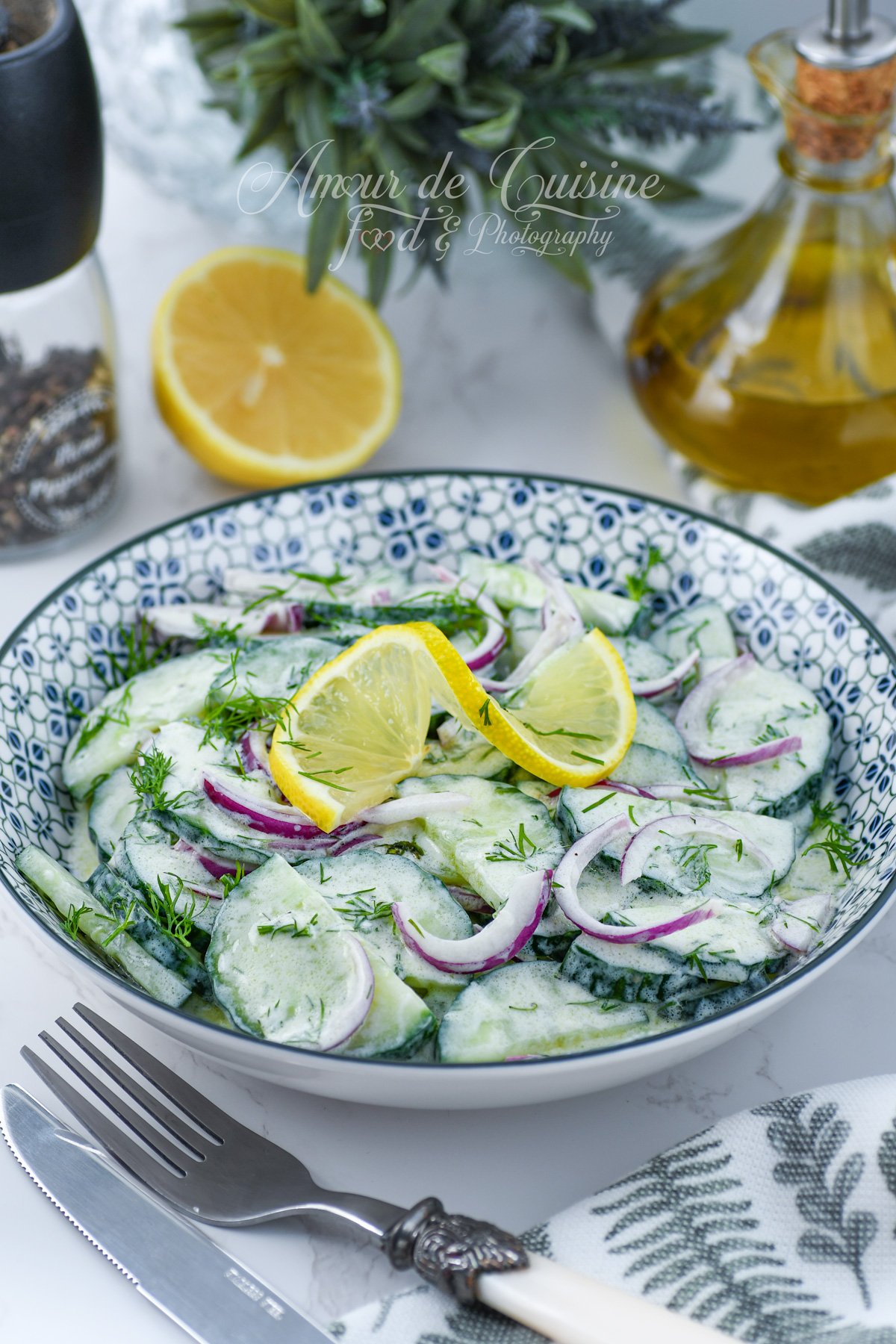 Bol de salade de concombre au yaourt, aneth et oignon rouge, garni de quartiers de citron, sur un plan de travail clair avec huile d’olive et moulin à poivre en arrière-plan.