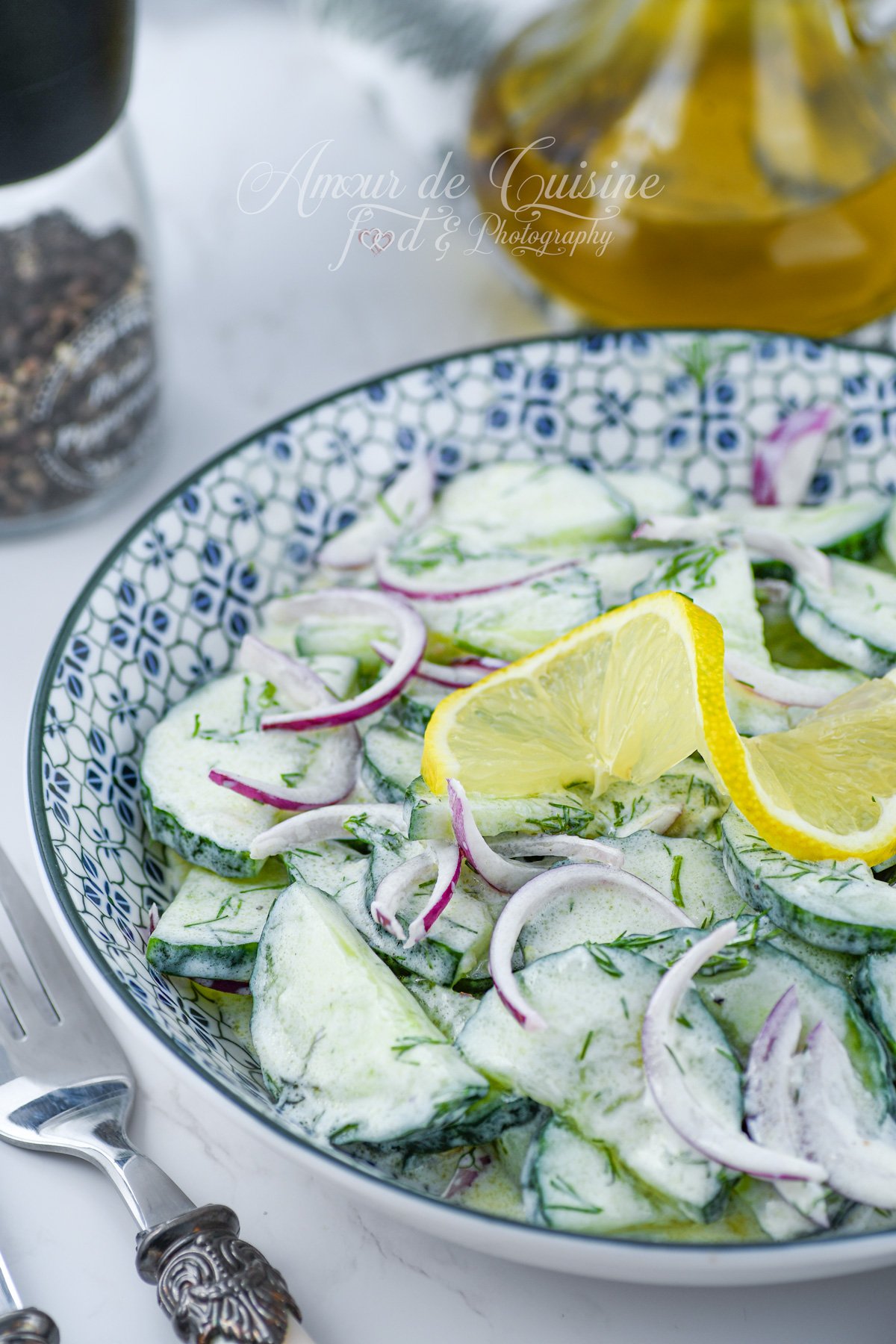 Gros plan sur une salade de concombre au yaourt et à l’aneth, avec oignon rouge émincé et quartiers de citron, dans un bol à motifs, avec huile d’olive et moulin à poivre en arrière-plan.