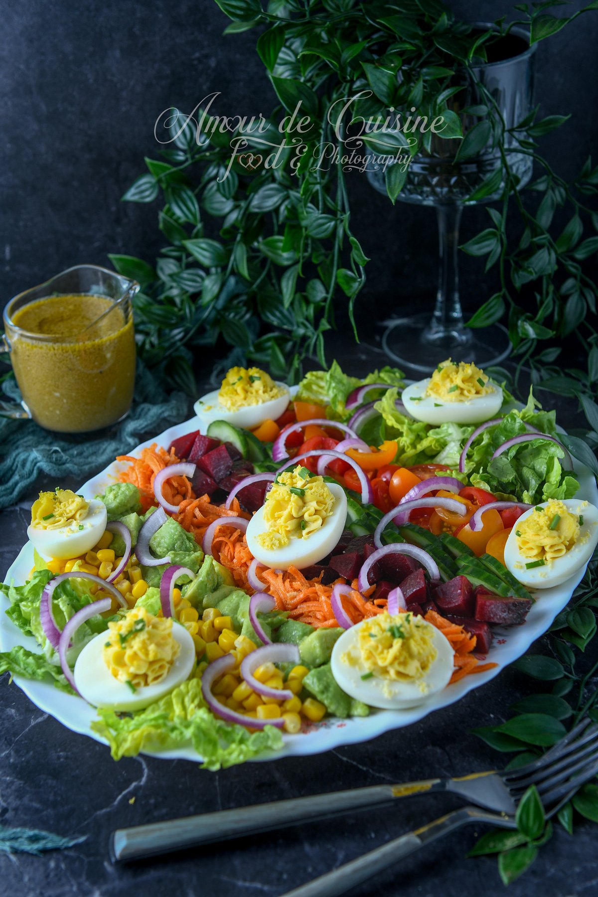 Grand plat de salade de crudités colorée avec œufs mimosa, carottes râpées, betteraves en dés, concombre, maïs, tomates cerises, oignon rouge et laitue, avec vinaigrette dans un petit pichet.