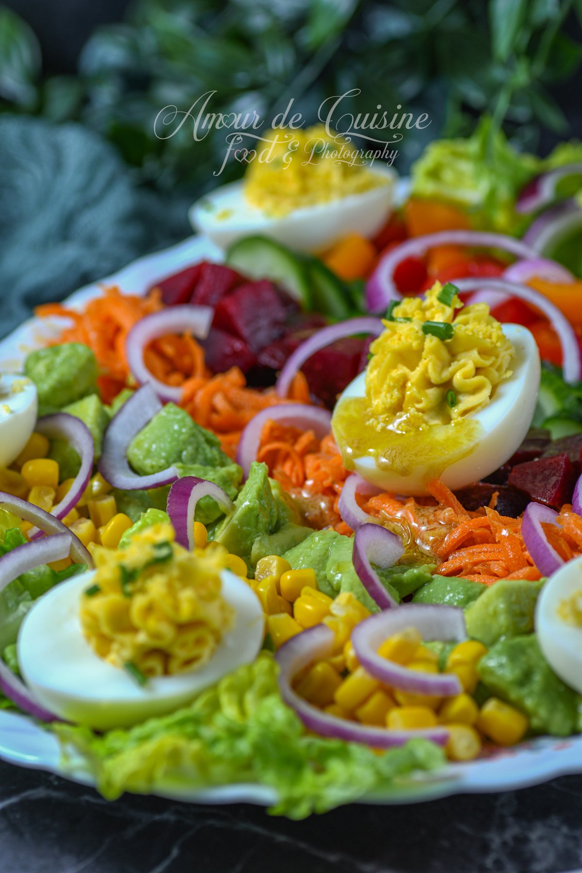 Gros plan sur une salade de crudités colorée avec œufs mimosa, maïs, avocat en cubes, carottes râpées, betteraves, concombre, tomates et rondelles d’oignon rouge, nappée d’un filet de vinaigrette.