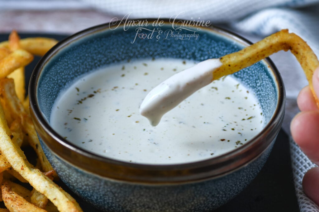 Gros plan d’une frite trempée dans une sauce ranch maison crémeuse aux herbes, dans un bol bleu texturé, avec des frites dorées en arrière-plan.