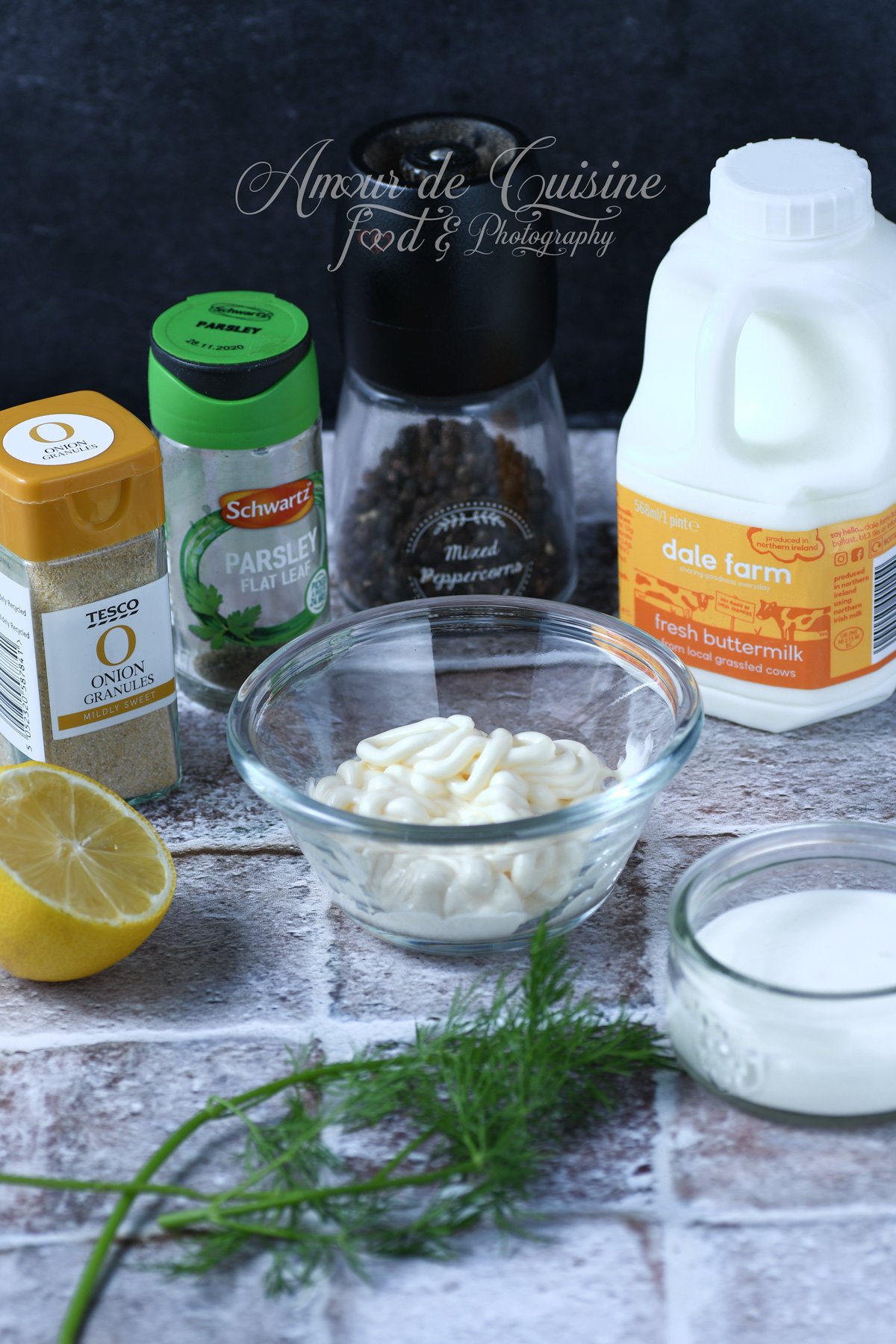 Ingrédients pour sauce ranch maison : mayonnaise en bol, babeurre, crème sure, citron, aneth frais, persil séché, poudre d’oignon et moulin à poivre sur un plan de travail.