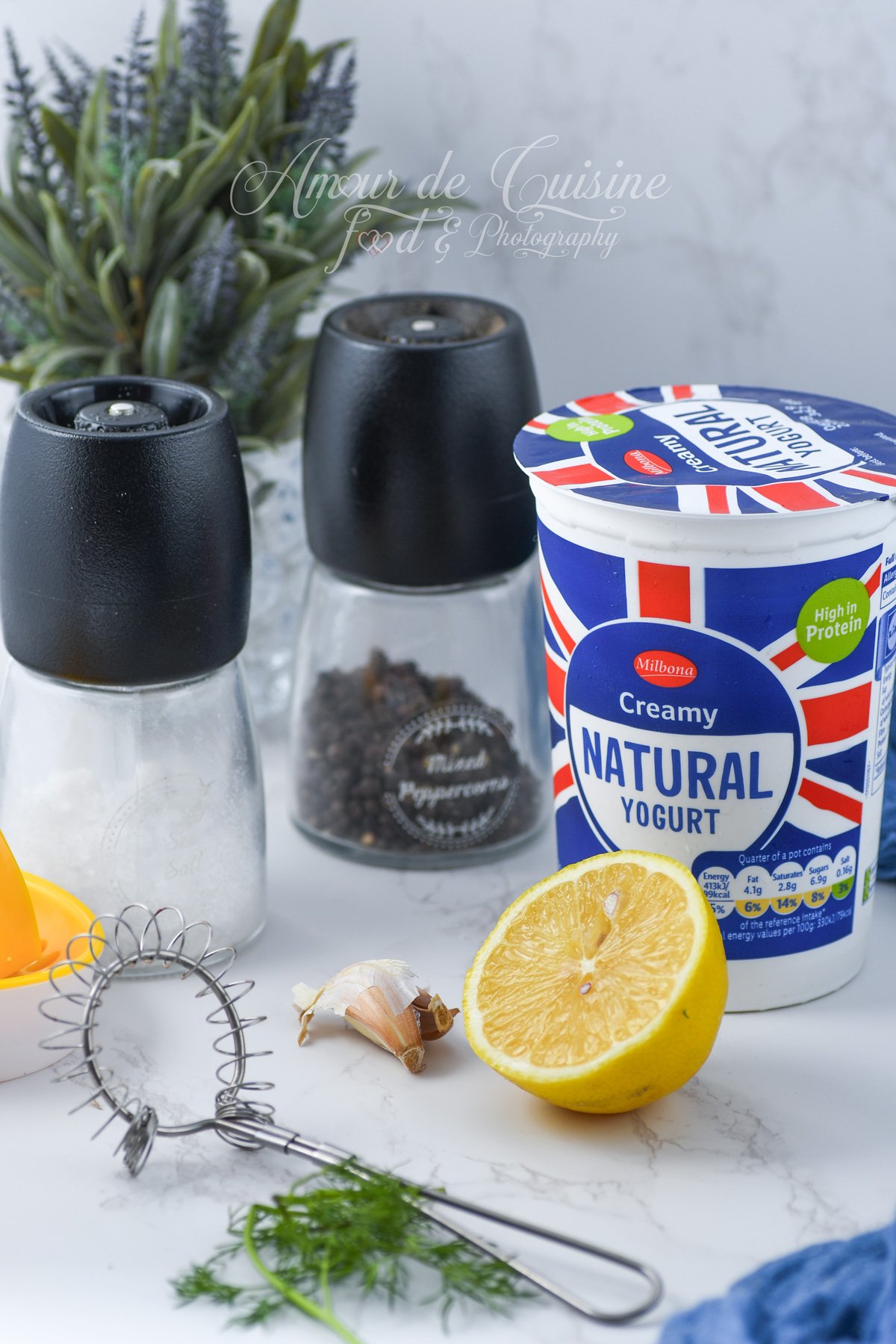 Ingrédients pour vinaigrette au yaourt : pot de yaourt nature, demi-citron, gousse d’ail, poivrier, salière et fouet spiralé sur plan de travail clair.