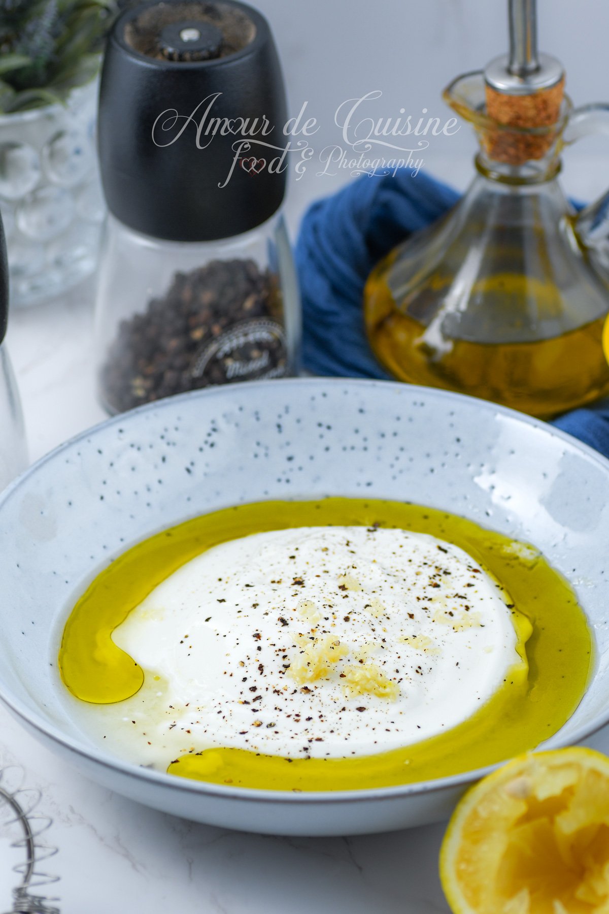Bol avec yaourt nature, huile d’olive, ail râpé et poivre avant mélange, pour une vinaigrette au yaourt ; poivrier et bouteille d’huile en arrière-plan.