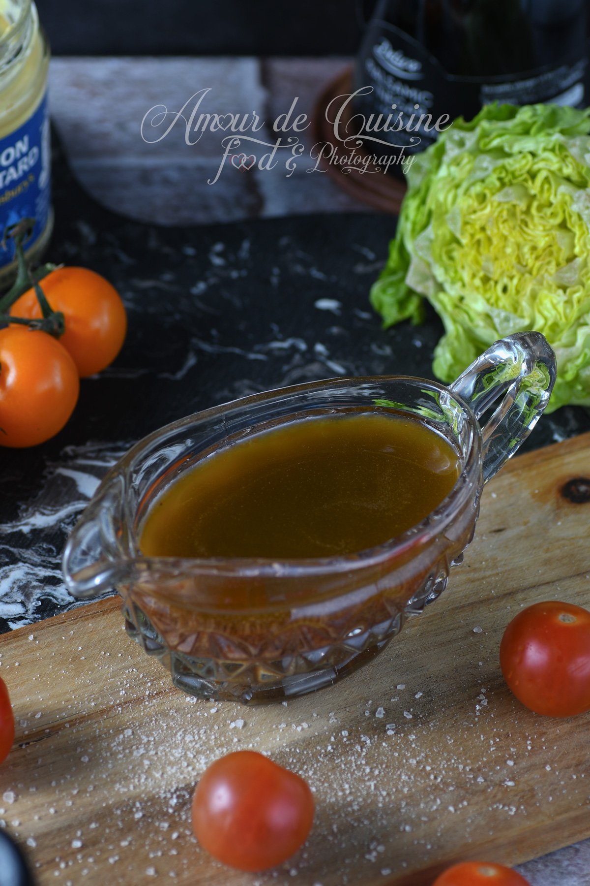 Vinaigrette balsamique à l’érable dans une saucière en verre sur une planche en bois, entourée de tomates cerises, salade verte et bouteille de vinaigre en arrière-plan.