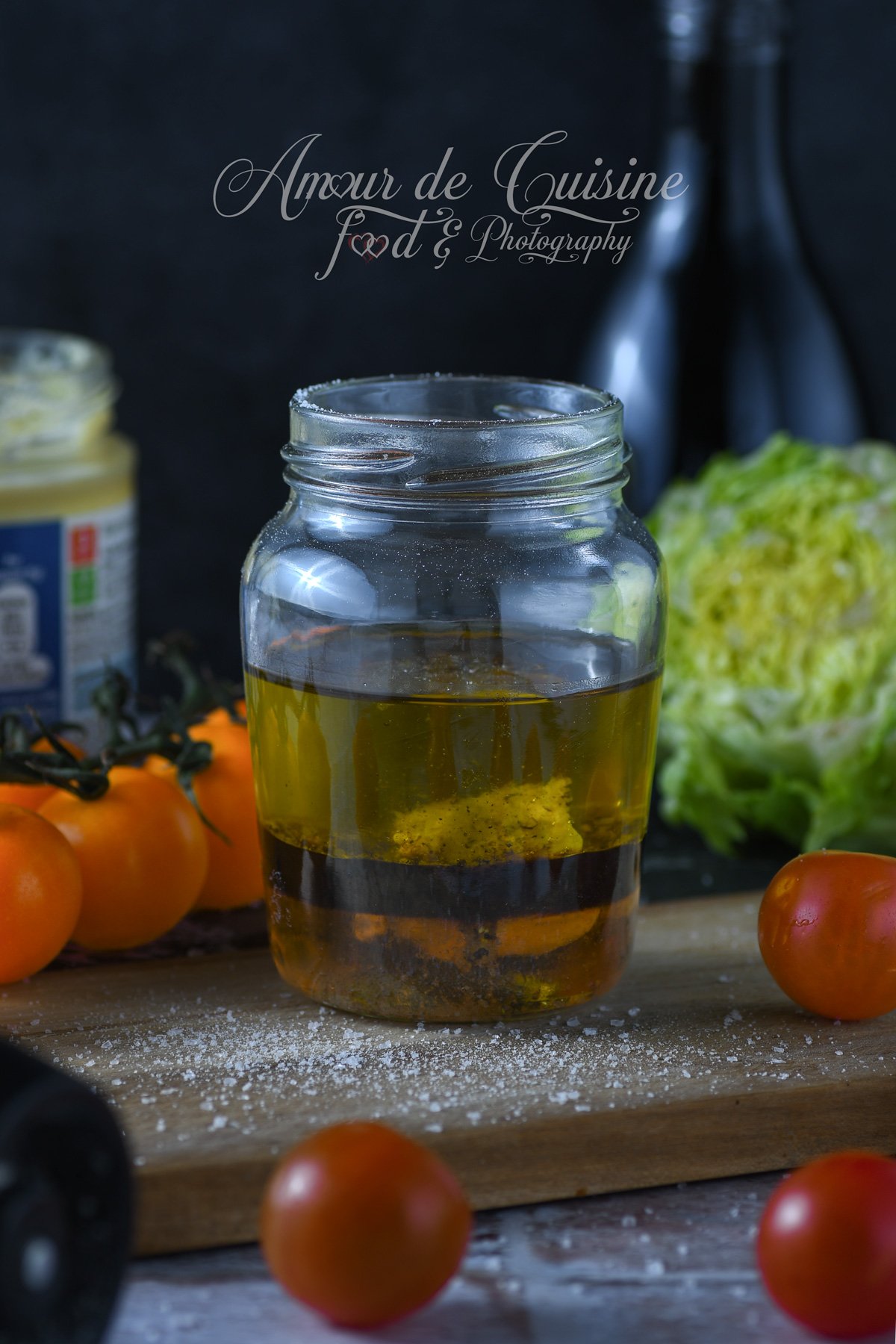 Vinaigrette balsamique à l’érable en cours de préparation dans un bocal, avec les couches d’huile, vinaigre et moutarde visibles, posée sur une planche avec tomates cerises et salade verte en arrière-plan.