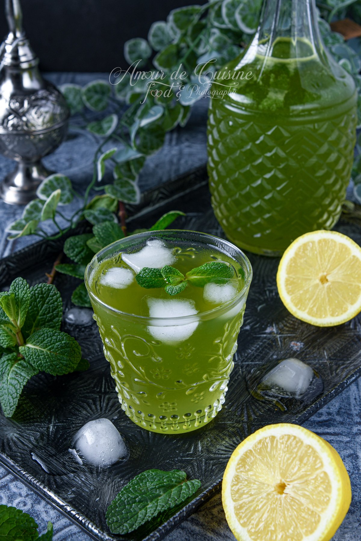 Verre de citronnade verte à la menthe avec glaçons, posé sur un plateau sombre, entouré de feuilles de menthe et de demi-citrons, avec une grande carafe de citronnade en arrière-plan.