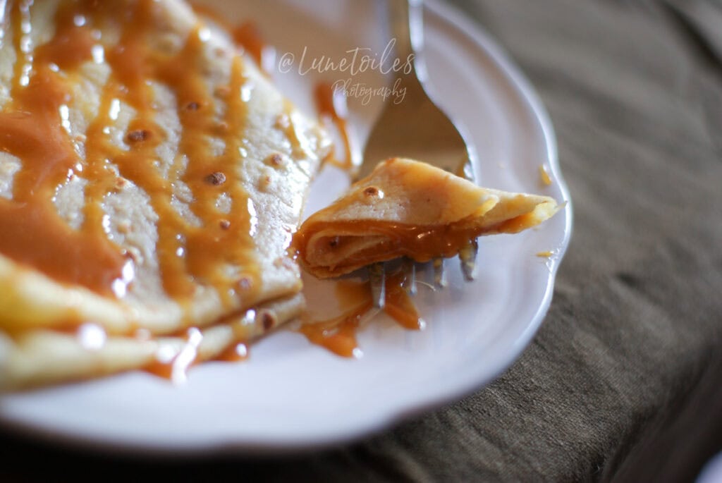Gros plan d’une crêpe nappée de caramel au beurre salé sur une assiette blanche, avec une bouchée roulée sur la fourchette et du caramel qui coule.