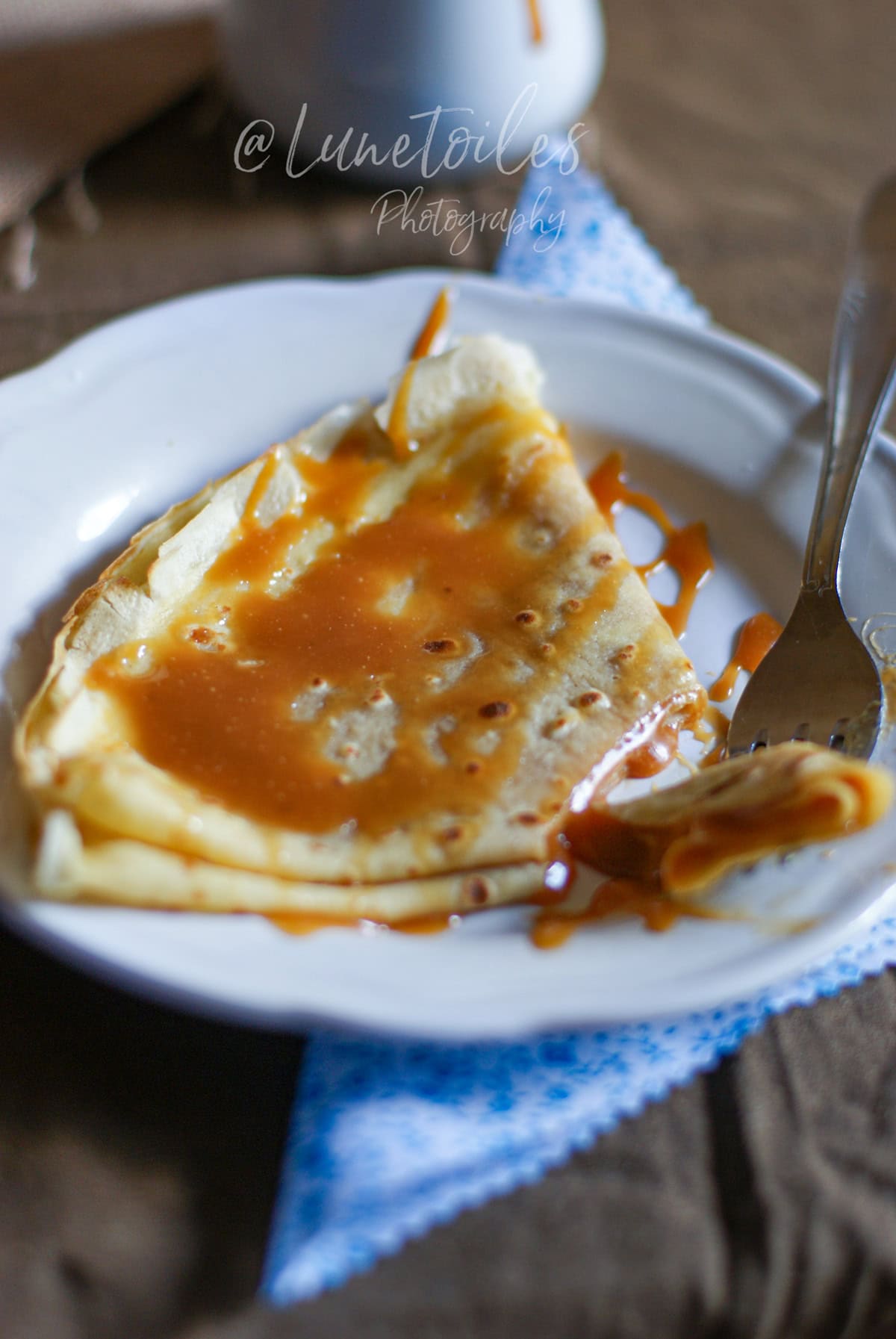 Crêpe pliée servie sur une assiette blanche, nappée de caramel au beurre salé, avec une fourchette tenant une bouchée, sur un torchon bleu en arrière-plan.