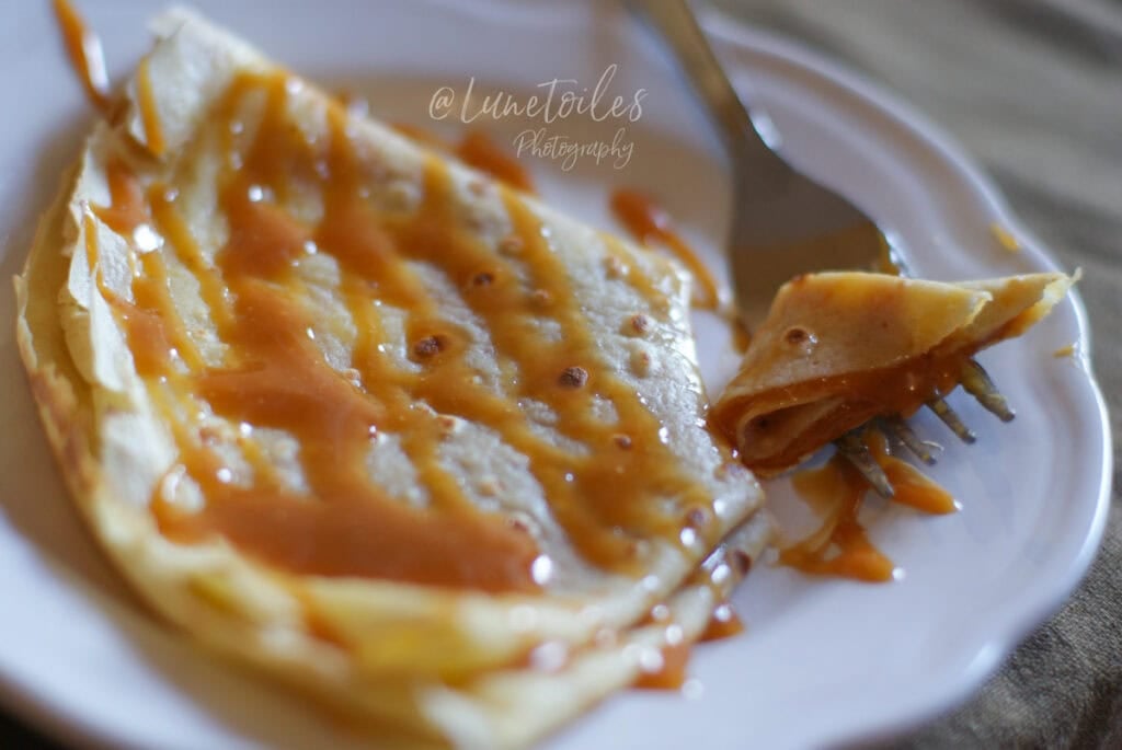 Crêpe pliée et nappée de caramel au beurre salé sur une assiette blanche, avec une bouchée roulée sur la fourchette et un caramel brillant qui nappe la surface.