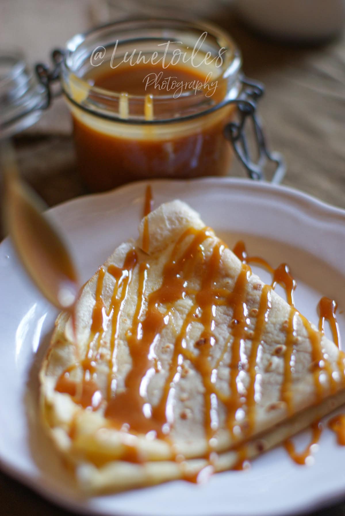 Crêpe pliée en triangle sur une assiette blanche, nappée de caramel au beurre salé en filet, avec un pot de caramel en arrière-plan légèrement flou.