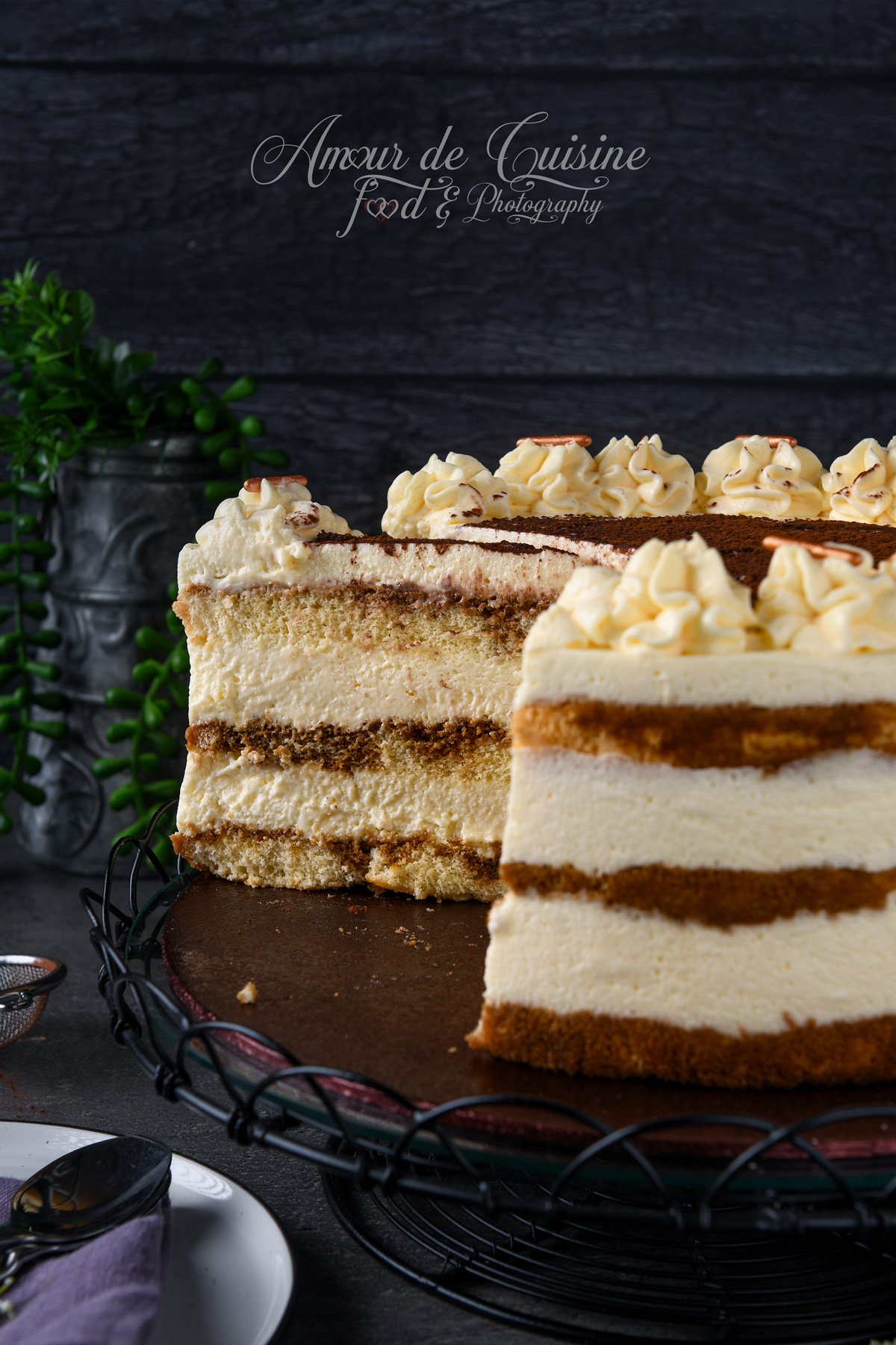 Gâteau tiramisu façon layer cake entamé sur un plateau, avec une grande part découpée au premier plan, montrant les couches de génoise imbibée au café et de mousse mascarpone, surmonté de rosaces de crème et de cacao.