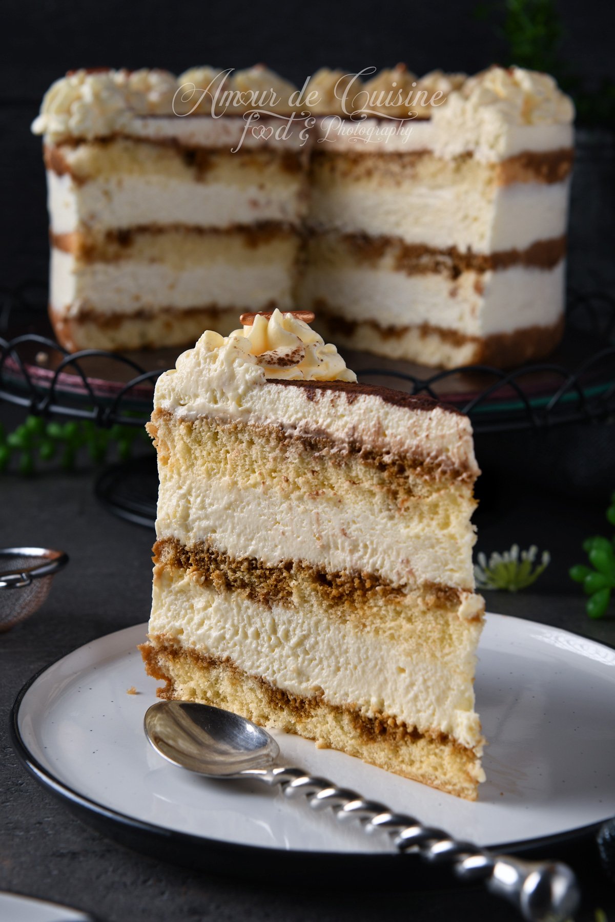 Part de gâteau tiramisu façon layer cake sur une assiette, montrant les couches de génoise imbibée au café et de mousse mascarpone, avec une rosace de crème sur le dessus ; le gâteau entier apparaît flou en arrière-plan.