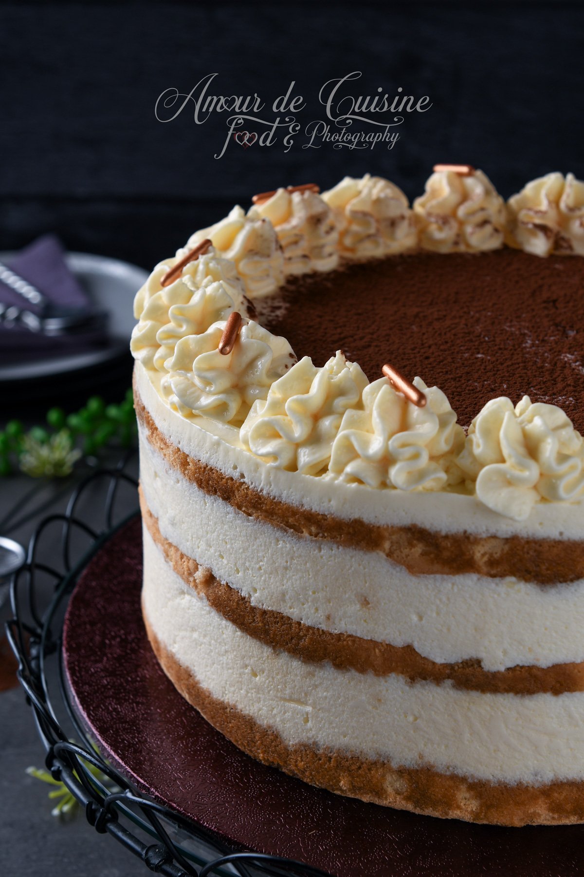 Gros plan sur un gâteau tiramisu façon layer cake, montrant les couches de génoise imbibée au café et de mousse mascarpone, avec une couronne de rosaces de crème et un centre saupoudré de cacao.