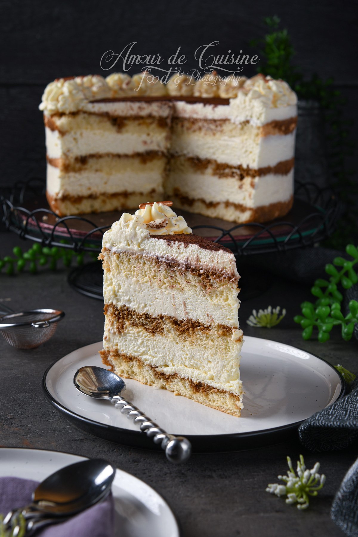 Part de gâteau tiramisu façon layer cake (génoise imbibée au café et mousse mascarpone), posée sur une assiette avec une cuillère, avec le gâteau entier en arrière-plan, décoré de cacao et de rosaces de crème.