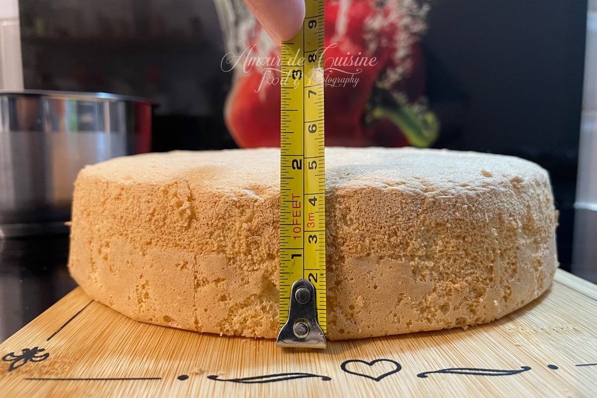 Génoise entière posée sur une planche, mesurée en hauteur avec un mètre ruban tenu à la main, pour montrer l’épaisseur du gâteau avant de le découper en disques.
