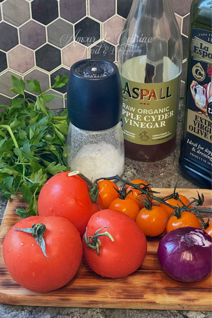 Ingrédients pour salade de tomates : tomates rouges et tomates cerises orange, oignon rouge, persil frais, sel, vinaigre de cidre et huile d’olive sur planche en bois.