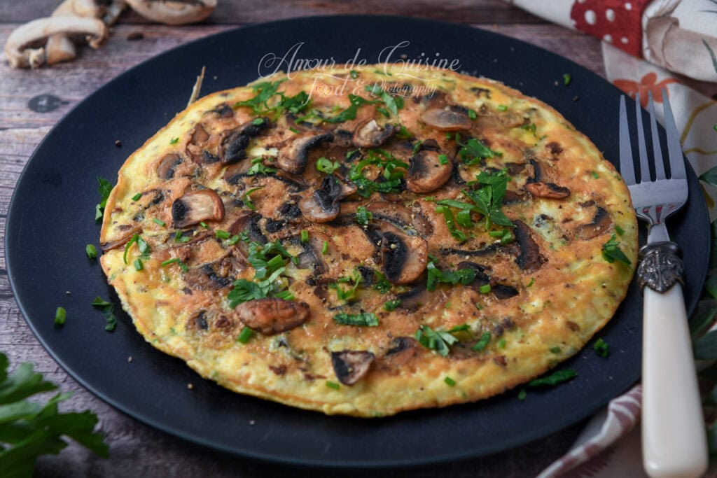 Omelette aux champignons de Paris dorée et moelleuse, garnie de persil et ciboulette, servie entière sur une grande assiette noire avec une fourchette à manche blanc.