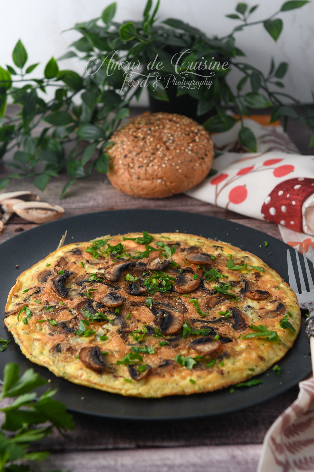 Omelette aux champignons de Paris parsemée de persil et ciboulette, servie sur une assiette noire, avec un pain aux graines et des plantes en arrière-plan.