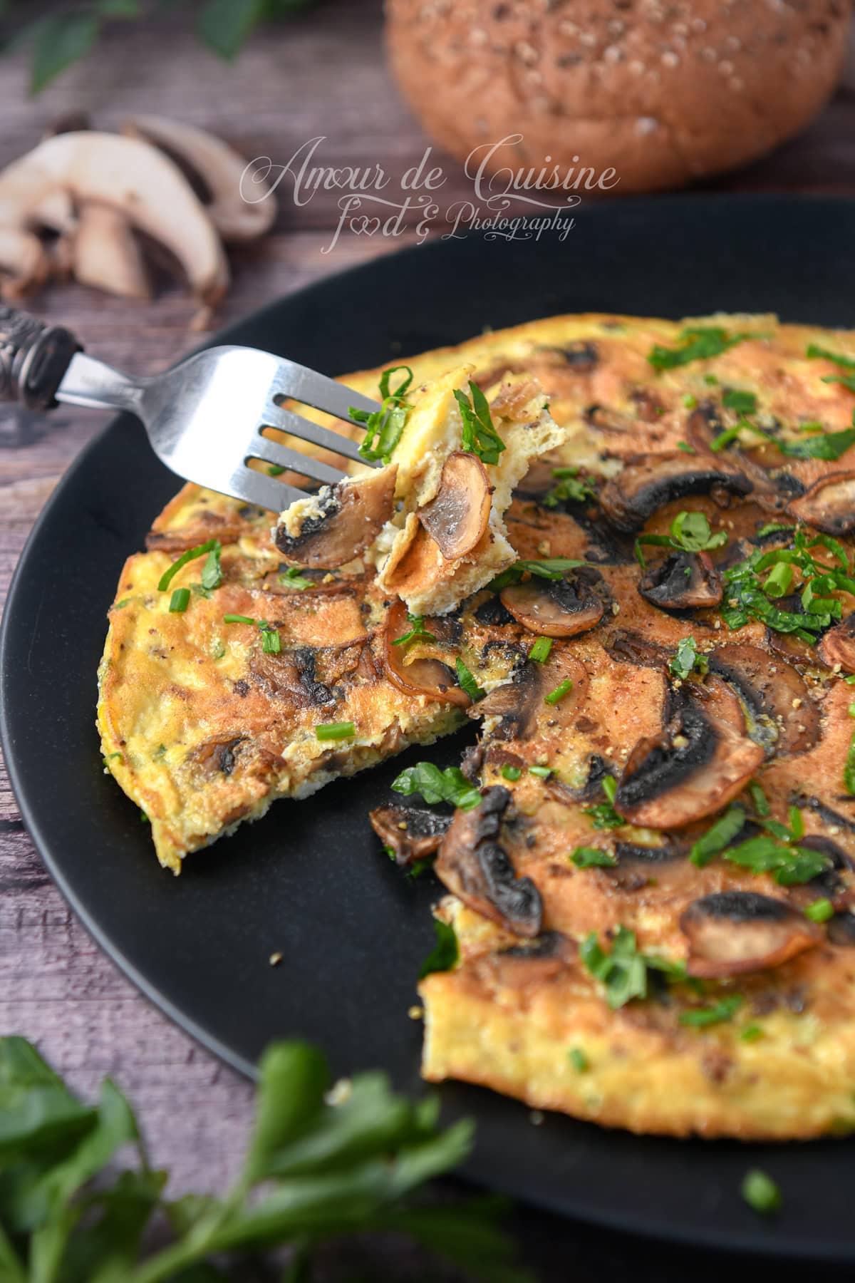 Part d’omelette aux champignons de Paris, parsemée de persil et ciboulette, servie sur une assiette noire avec une fourchette soulevant une bouchée.