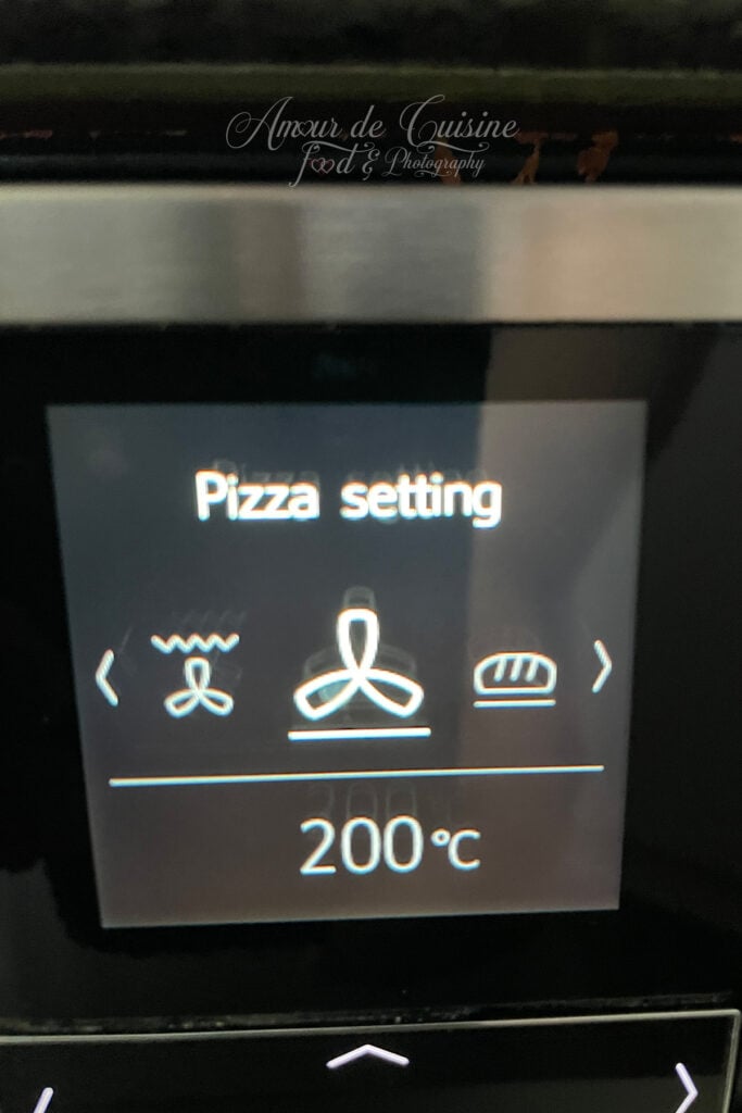 : Écran d’un four affichant le mode “Pizza setting” à 200 °C, avec icône ventilateur et chaleur par le bas, ainsi qu’un symbole de pain sur le panneau tactile.