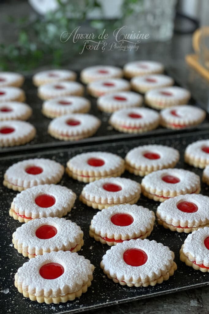 Sablés à la confiture de fraises alignés sur plaque, saupoudrés de sucre glace, avec centre rouge brillant et texture fondante.