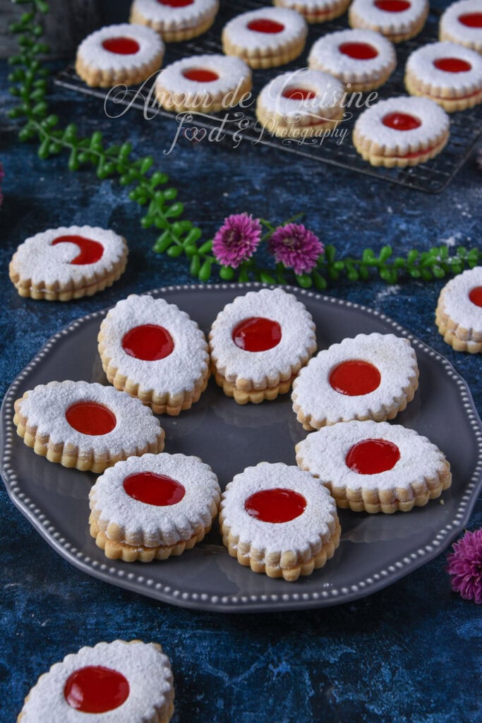 Sablés à la confiture de fraises dressés sur un plateau gris, saupoudrés de sucre glace, avec biscuits assortis en arrière-plan.