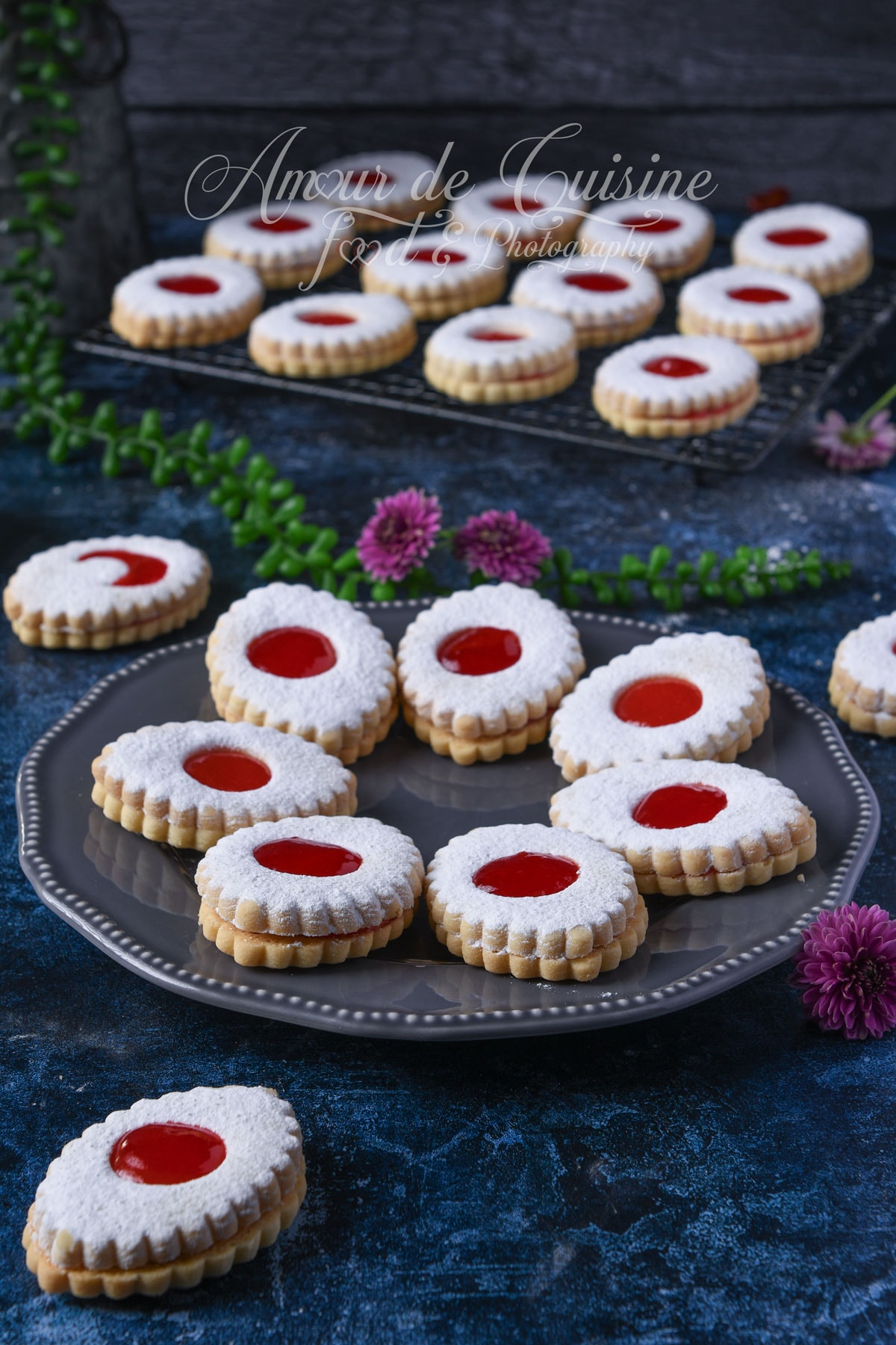 Sablés à la confiture de fraises saupoudrés de sucre glace, présentés sur un plateau gris avec d’autres biscuits en arrière-plan.