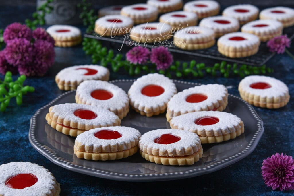Sablés à la confiture de fraises présentés sur un plateau gris, saupoudrés de sucre glace, avec centre rouge brillant et biscuits en arrière-plan.