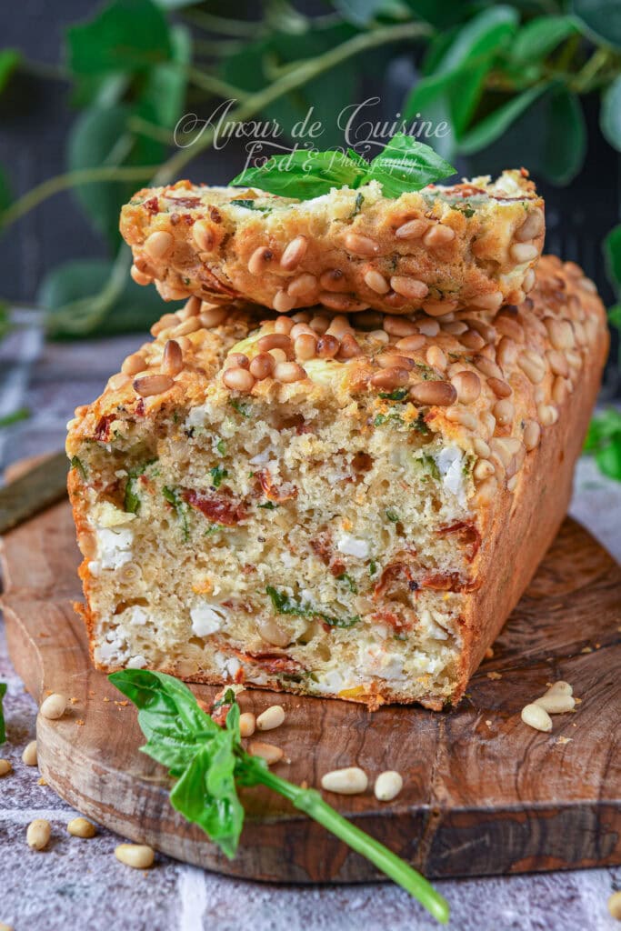 Cake salé aux tomates séchées, feta, basilic et pignons de pin, avec une tranche posée sur le dessus pour montrer la mie garnie.