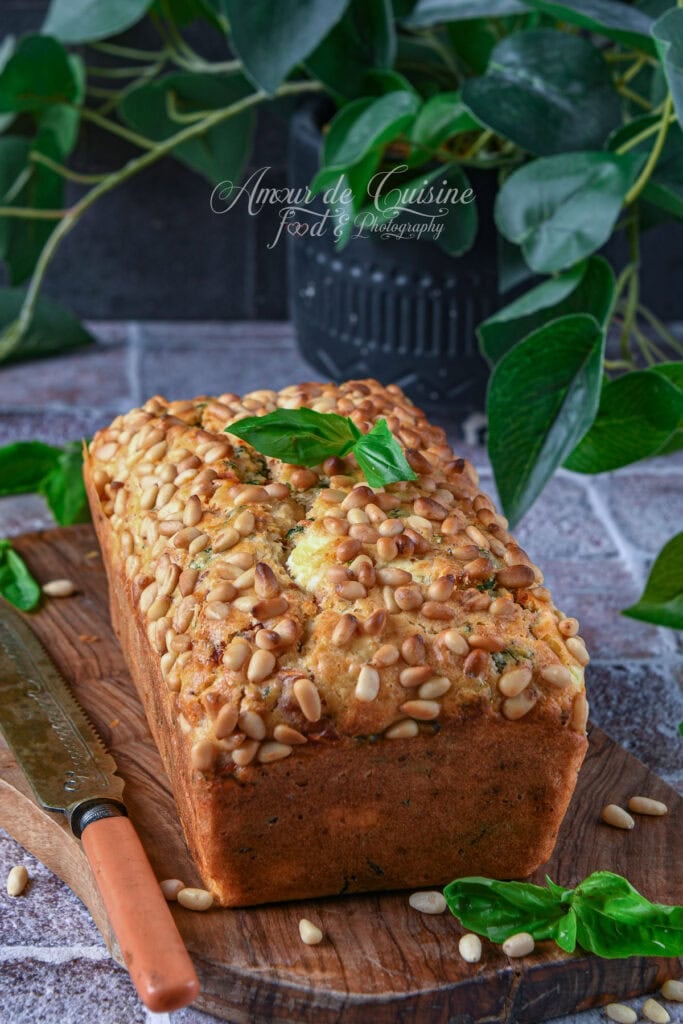 Cake salé doré aux tomates séchées, feta, basilic et pignons de pin, présenté entier sur une planche en bois.