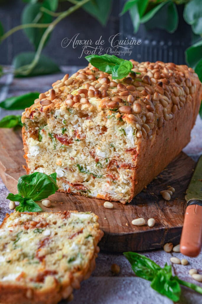 Cake salé aux tomates séchées, feta, basilic frais et pignons de pin, coupé en tranches sur une planche en bois.