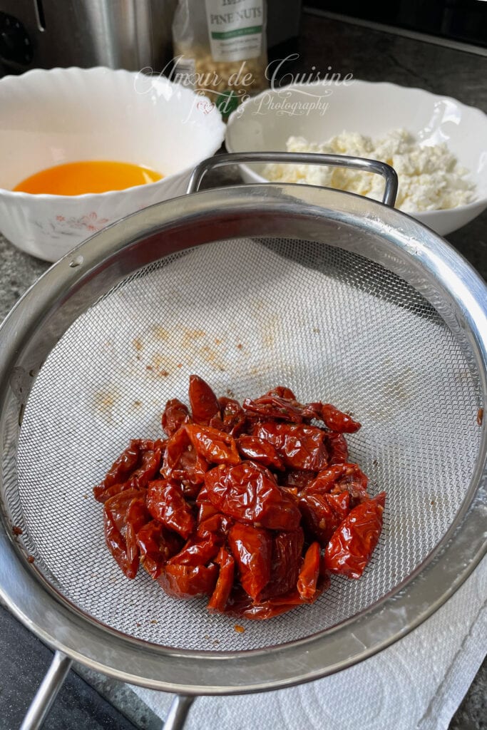 Tomates séchées confites égouttées dans une passoire, avec feta émiettée et œufs préparés pour un cake salé.